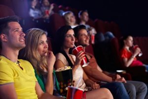 Young people sitting in multiplex movie theater, watching movie, eating popcorn.